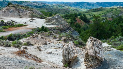 A rocky landscape with large boulders and rolling hills in the background, featuring green vegetation and a cloudy sky.