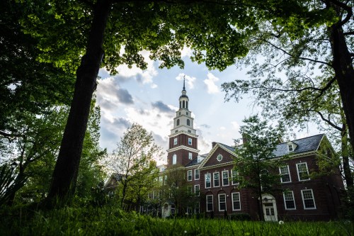A historic brick building with a clock tower, surrounded by lush green trees and a cloudy sky.