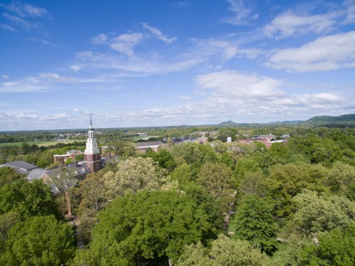 Aerial view of a lush green campus with buildings and a clock tower under a blue sky with scattered clouds.
