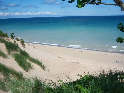 A serene beach scene with soft sand dunes, green grass, and calm blue waters under a partly cloudy sky.