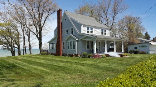 A charming gray house with a porch, surrounded by green grass and trees, near a lake on a sunny day.