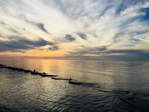 A serene sunset over calm waters, with clouds reflecting colors in the sky and silhouettes of boats on the horizon.