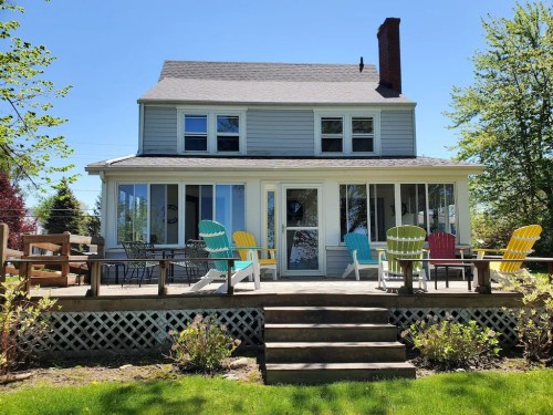 A charming two-story house with a porch, surrounded by greenery and colorful chairs on a sunny day.