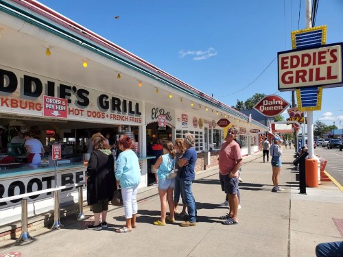A group of people waiting in line outside Eddie's Grill, with signs for hot dogs and Dairy Queen in the background.