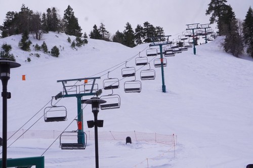 Ski lift chairs ascend a snowy slope, surrounded by trees under a cloudy sky.