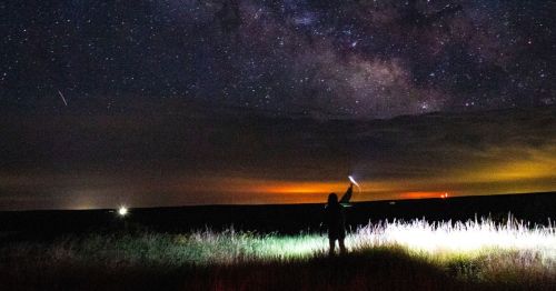 A silhouette of a person holding a light against a starry night sky, with distant lights and clouds in the background.