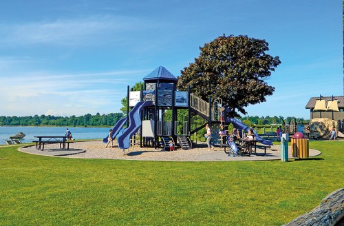 A playground with slides and climbing structures near a lake, surrounded by green grass and trees on a sunny day.