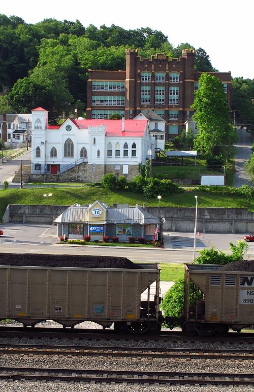 A view of a town with a church, a school building, and a train with coal cars in the foreground. Green hills in the background.
