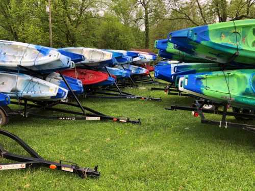 A stack of colorful kayaks on trailers, resting on green grass in a park-like setting.