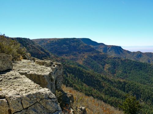A rocky cliff overlooks a lush, green mountain range under a clear blue sky.