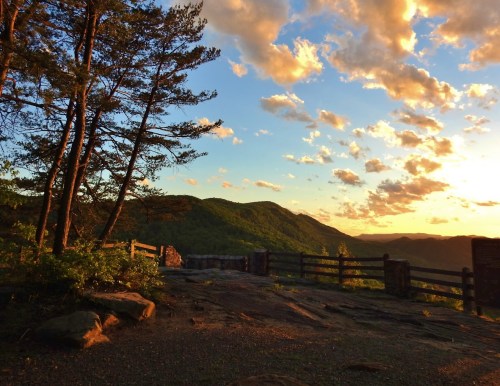 A scenic view of mountains at sunset, with trees and a wooden fence in the foreground under a colorful sky.