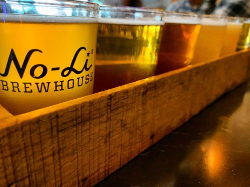A wooden flight tray holds several glasses of different beers, showcasing a variety of colors and styles.