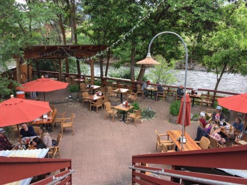 Outdoor dining area with wooden tables, red umbrellas, and a river view, surrounded by greenery and patrons enjoying meals.