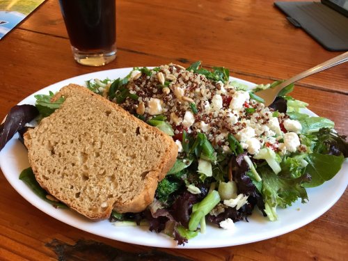 A fresh salad with greens, quinoa, feta cheese, and a slice of brown bread on a white plate, with a drink in the background.