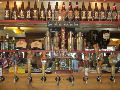 A bar shelf displaying various beer bottles, trophies, and mugs, with a sign reading "Good Food, Good Friends, Good Times."