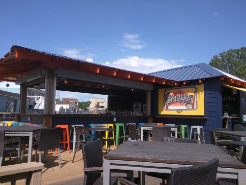Outdoor bar with colorful stools and tables, featuring a sign that reads "Bridges," under a clear blue sky.