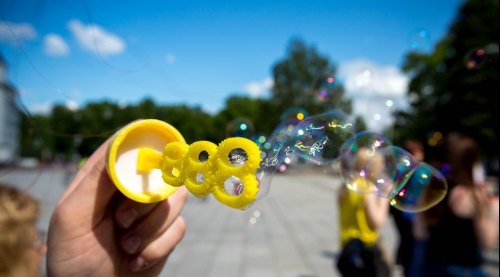 A hand holds a yellow bubble wand, creating colorful bubbles against a bright blue sky and blurred background.