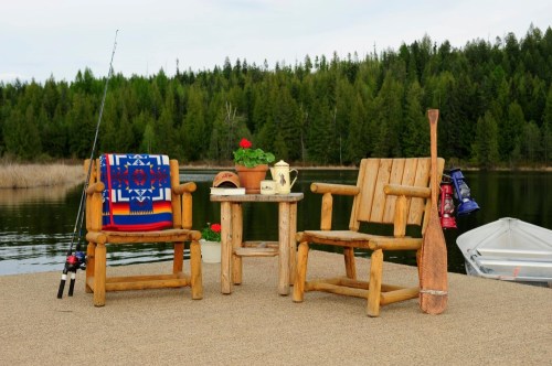 Two rustic wooden chairs on a dock by a lake, with a fishing rod, a potted plant, and a decorative blanket.