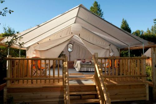 A spacious canvas tent with a wooden deck, featuring a dining area and surrounded by trees in a sunny outdoor setting.
