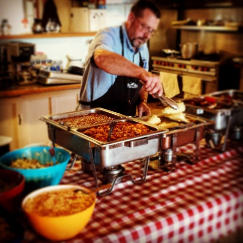 A man serves food from a buffet with various dishes in colorful bowls on a checkered tablecloth.