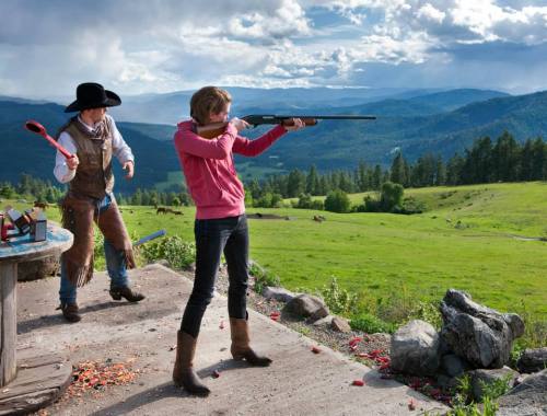 A woman aims a shotgun at a distant target while a man stands nearby, with a scenic mountain landscape in the background.