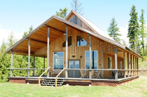 A wooden cabin with a large porch, surrounded by trees and blue sky, showcasing a rustic, inviting design.