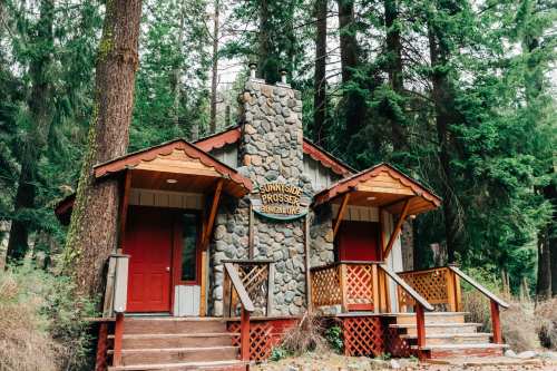 A rustic cabin with a stone chimney, surrounded by tall trees, featuring two red doors and a sign reading "Sunnyside Prosser Bungalows."