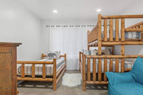 Cozy bedroom with two sets of wooden bunk beds, a blue chair, and a soft rug, featuring light curtains.