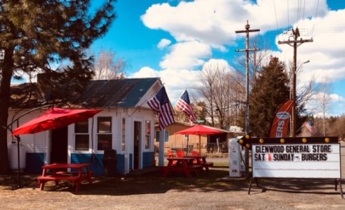 A small general store with red umbrellas, picnic tables, and American flags, featuring a sign for burgers on weekends.