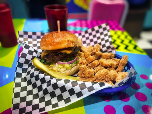 A burger with lettuce, onion, and pickles served with crispy fried shrimp on a colorful checkered table.