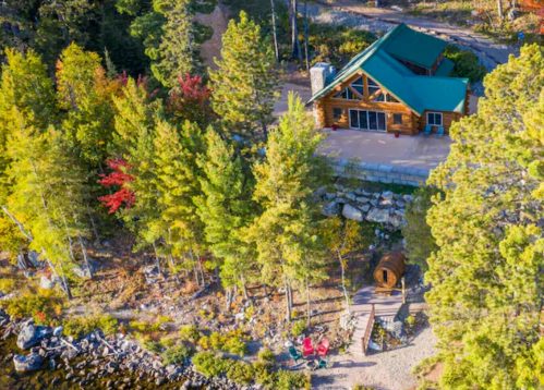 Aerial view of a cozy cabin surrounded by trees near a lake, with a dock and colorful autumn foliage.