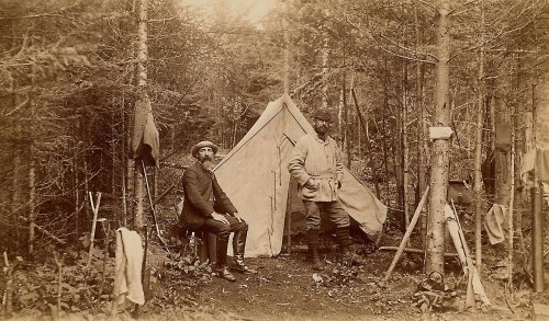 Two men stand beside a canvas tent in a forest, surrounded by trees and camping gear.