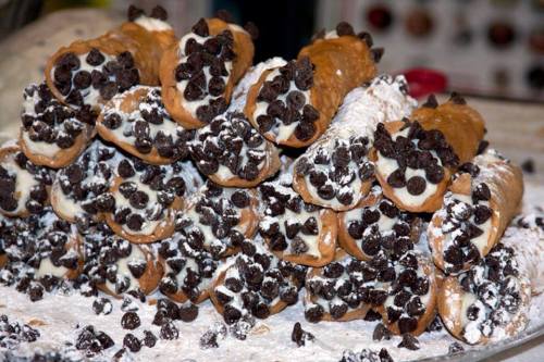 A stack of cannoli filled with cream and topped with chocolate chips, dusted with powdered sugar.