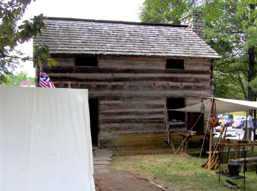 A rustic log cabin with a stone chimney, surrounded by tents and trees, set in a historical outdoor setting.