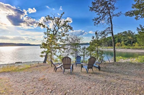 A serene lakeside scene with four Adirondack chairs arranged in a circle under a blue sky with scattered clouds.
