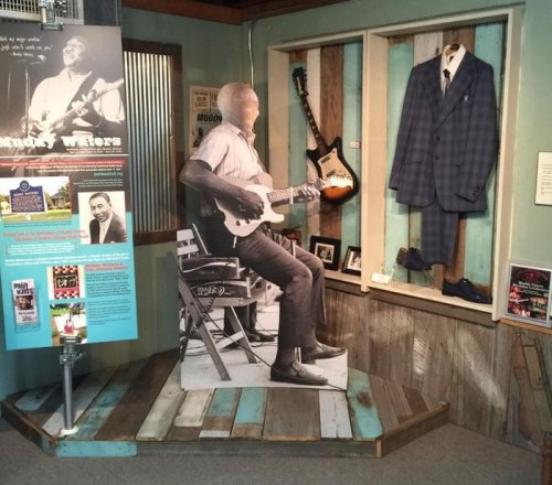 A life-size cutout of a guitarist in a museum exhibit, featuring a suit display and musical memorabilia.