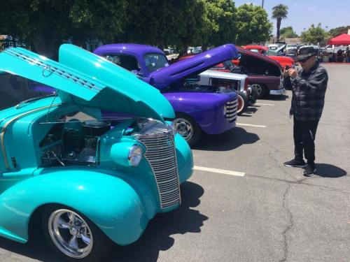 A person photographs classic cars with open hoods in a sunny parking lot, showcasing vibrant colors and vintage designs.