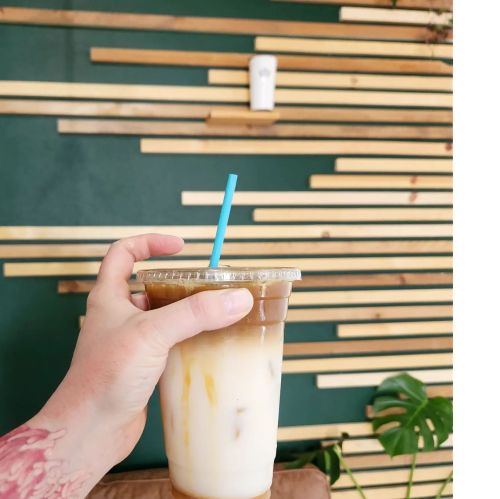 A hand holding a iced coffee drink with a blue straw in front of a wooden slat wall.