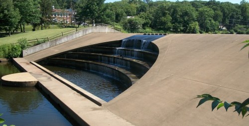 A modern concrete structure with cascading waterfalls flowing into a calm body of water, surrounded by greenery.