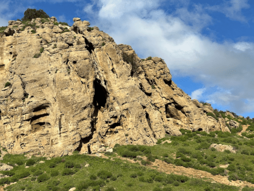 A rugged mountain with rocky cliffs and green vegetation under a partly cloudy blue sky.