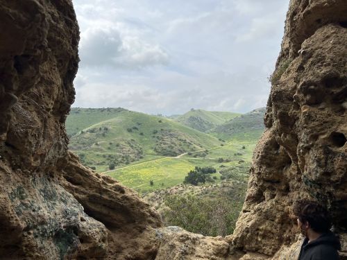 A person gazes out from a rocky opening, overlooking green hills and a cloudy sky.