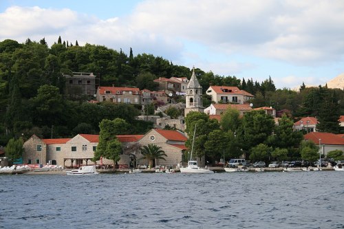A coastal village with stone buildings, red roofs, and lush greenery, set against a serene blue water backdrop.