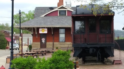 Historic train depot with a red train car attached, featuring outdoor seating and a sign for a restaurant.