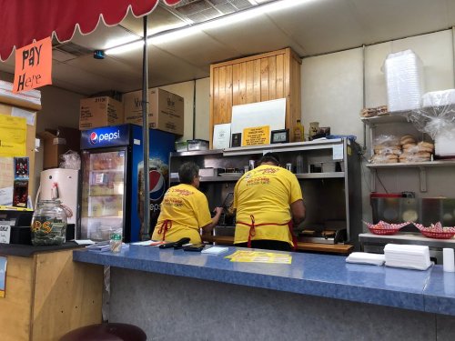 Two cooks in yellow shirts prepare food behind a counter in a small diner, with drinks and supplies visible.