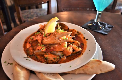 A plate of seafood pasta topped with crab, shrimp, and a lemon wedge, served with slices of bread and a blue cocktail.