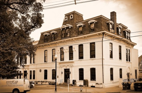 Historic building with a distinctive roof and large windows, surrounded by trees and a parked van. Sepia-toned image.