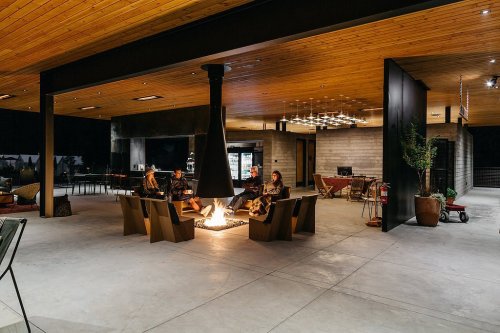 A modern outdoor space with a central fire pit, seating, and a wooden ceiling, featuring people gathered around the fire.