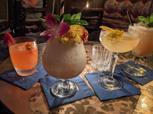 Three colorful cocktails with garnishes on a table, surrounded by decorative napkins and a cozy ambiance.