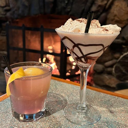 Two cocktails sit on a table in front of a cozy fireplace: a pink drink with a lemon slice and a chocolate dessert martini.