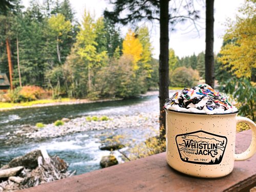 A cozy mug topped with whipped cream and sprinkles sits by a river, surrounded by autumn trees.